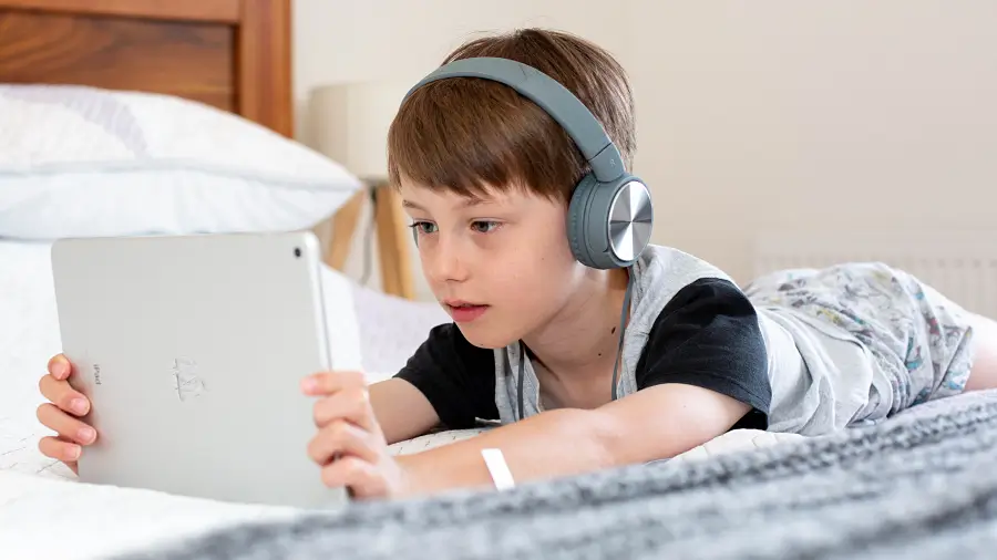 Kind liegt mit dem Tablet im Bett und hat Bluetooth-Headphone auf